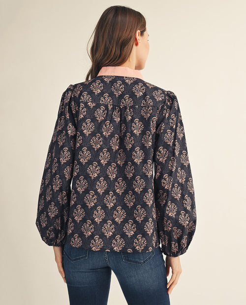 A woman viewed from the back wearing a long-sleeve blouse with a dark navy background and a repeating beige damask pattern, paired with blue jeans, against a solid light beige studio background.