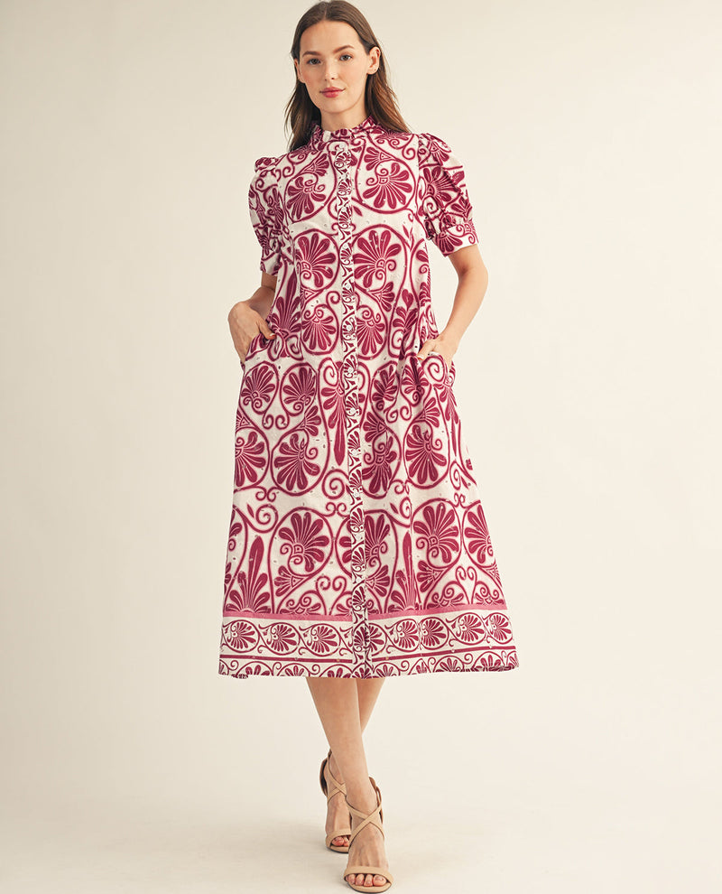 A woman modeling a short-sleeve midi dress with a pink and white floral block-print pattern, standing against a solid light beige background.