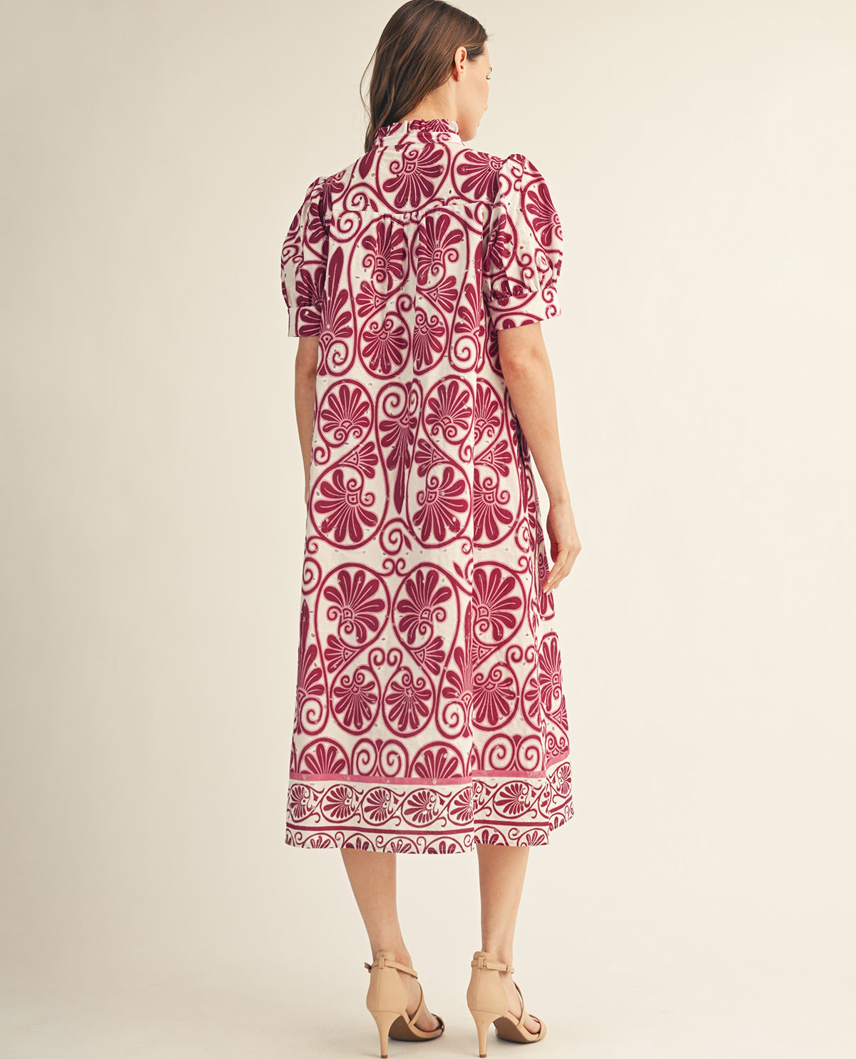 A woman viewed from the back wearing a short-sleeve midi dress with a bold magenta and cream floral and scroll pattern, paired with beige high-heeled sandals against a plain beige studio background.