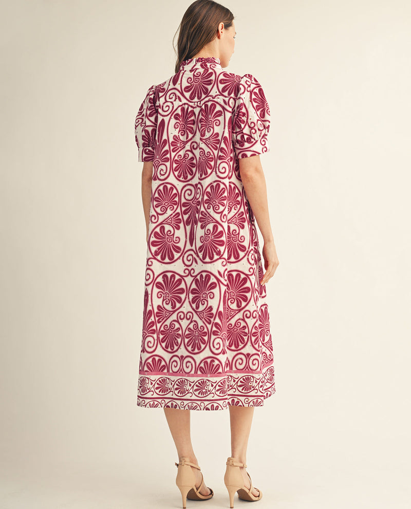 A woman viewed from the back wearing a short-sleeve midi dress with a bold magenta and cream floral and scroll pattern, paired with beige high-heeled sandals against a plain beige studio background.