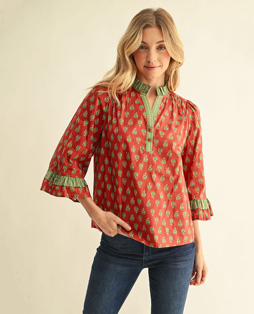 A woman modeling a red and green patterned blouse with 3/4 bell sleeves and green ruffled trim, paired with blue jeans, against a plain beige background.