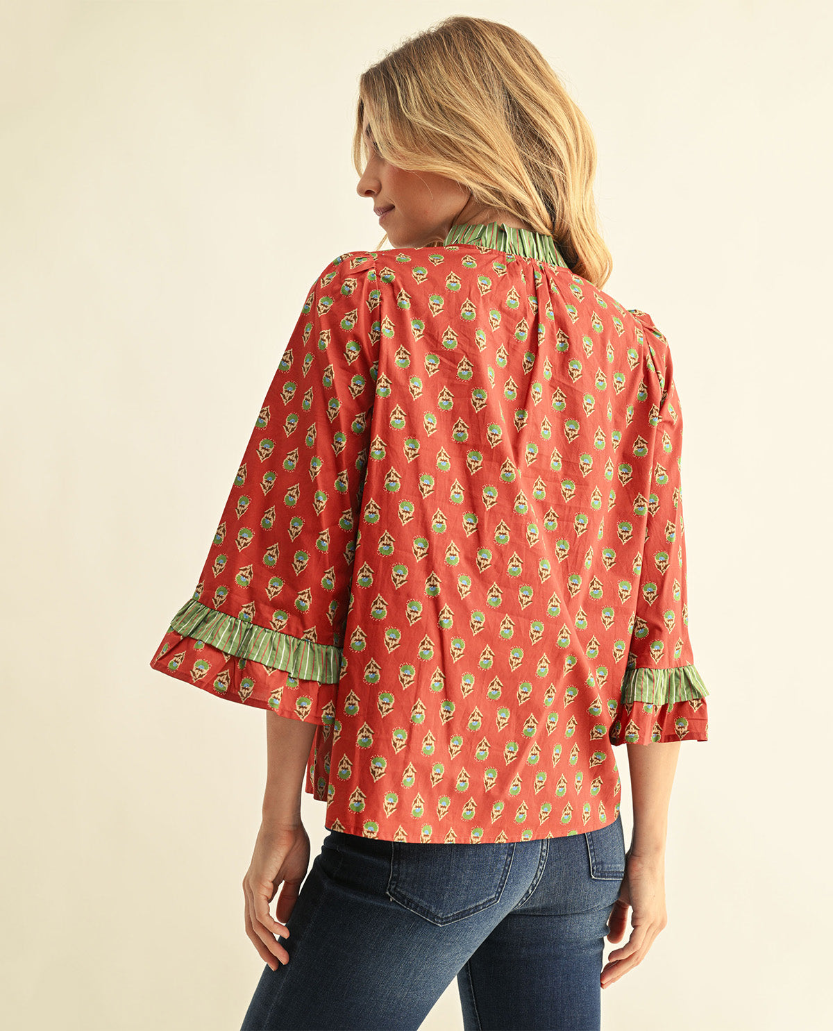 A woman shown from the back wearing a rust-orange blouse with a green and white floral print, ruffled trim at the collar and cuffs, and blue jeans against a plain cream background.