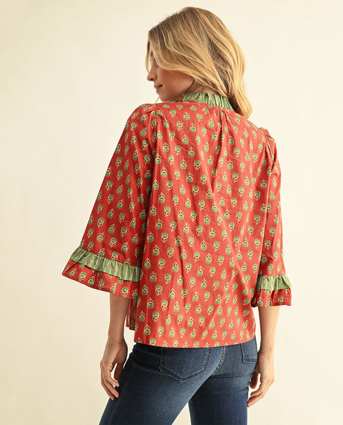A woman shown from the back wearing a rust-orange blouse with a green and white floral print, ruffled trim at the collar and cuffs, and blue jeans against a plain cream background.