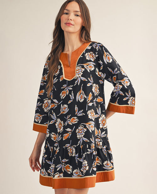 A woman modeling a black tunic dress with a white and orange floral pattern, featuring a contrasting orange V-neckline and orange-trimmed cuffs and hem, set against a plain beige background.