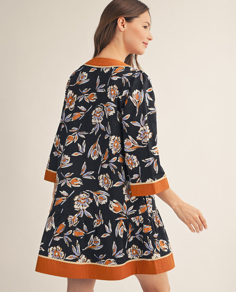 A woman wearing a black floral print dress with orange and white accents, shown from a side-back angle against a plain beige studio background.