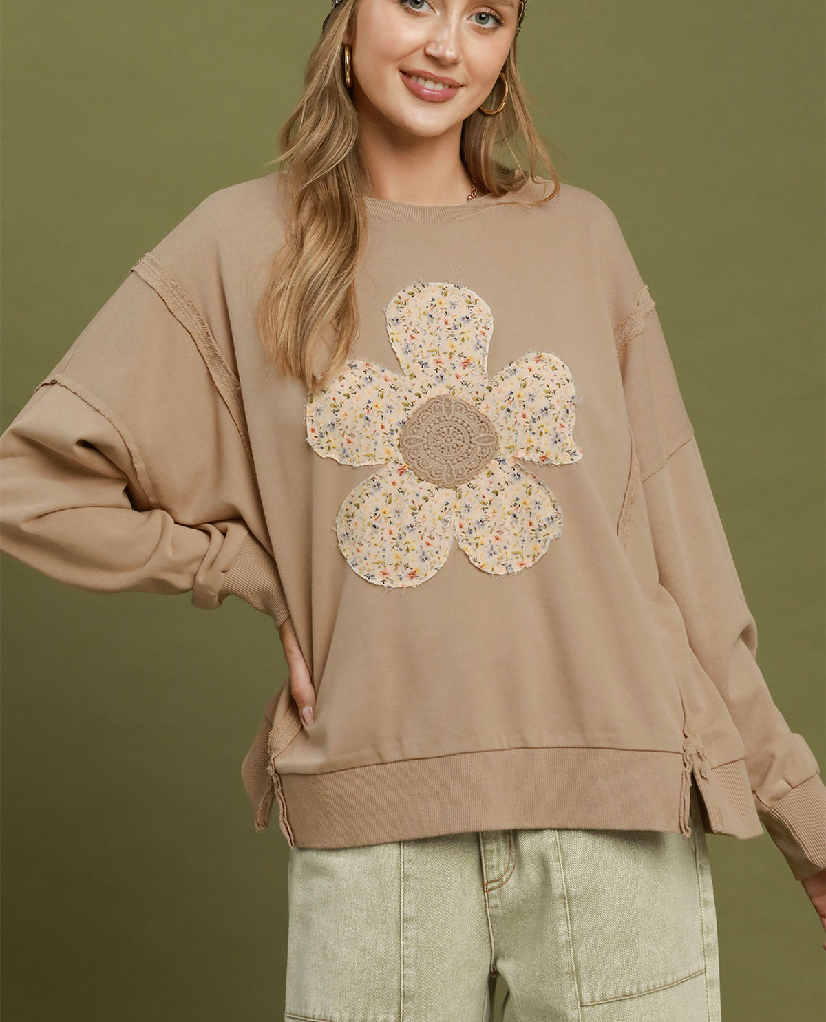 A woman wearing a beige oversized sweatshirt with a large floral applique on the front, paired with light green cargo pants, set against a solid olive green background.