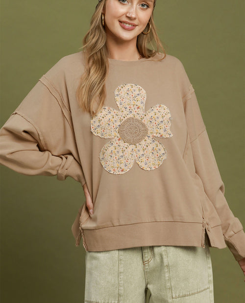 A woman wearing a beige oversized sweatshirt with a large floral applique on the front, paired with light green cargo pants, set against a solid olive green background.