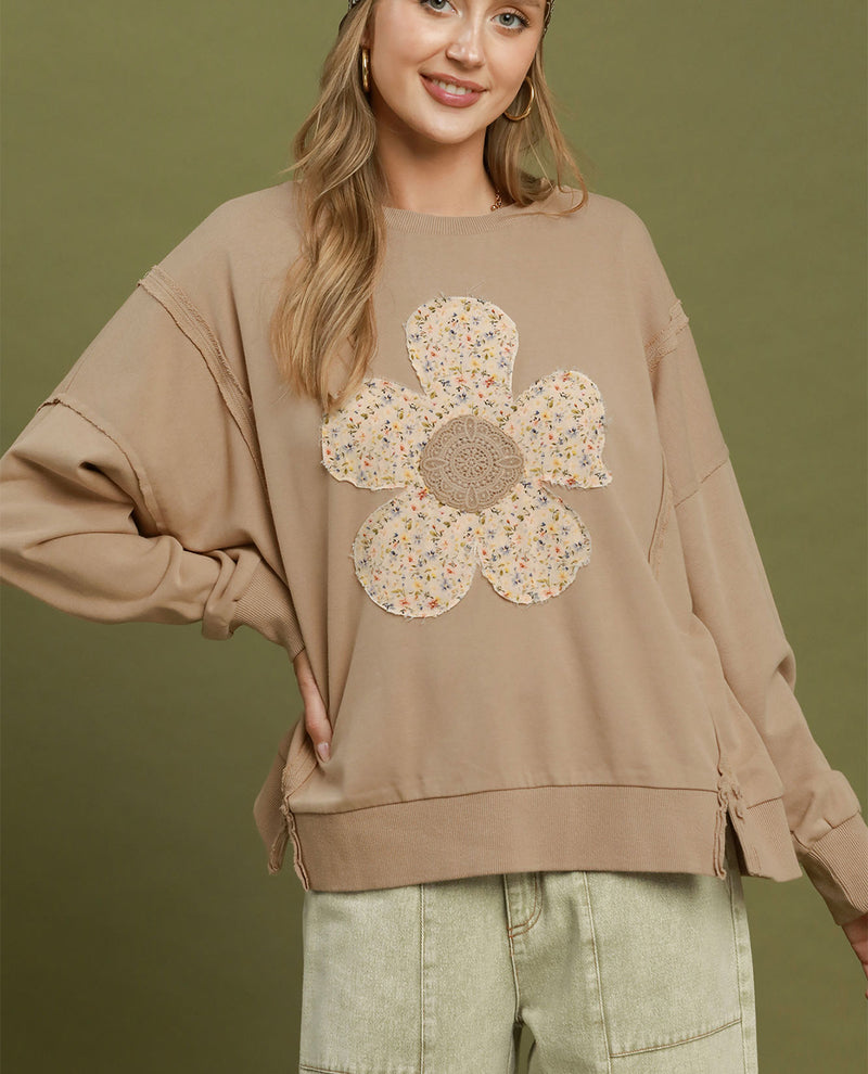 A woman wearing a beige oversized sweatshirt with a large floral applique on the front, paired with light green cargo pants, set against a solid olive green background.