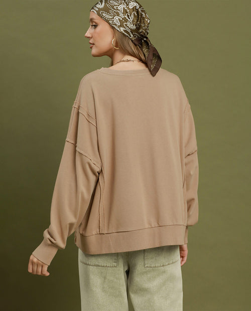 A woman shown from the back wearing an oversized taupe-beige long-sleeve sweatshirt and light olive pants, with a patterned headscarf against a solid olive green background.