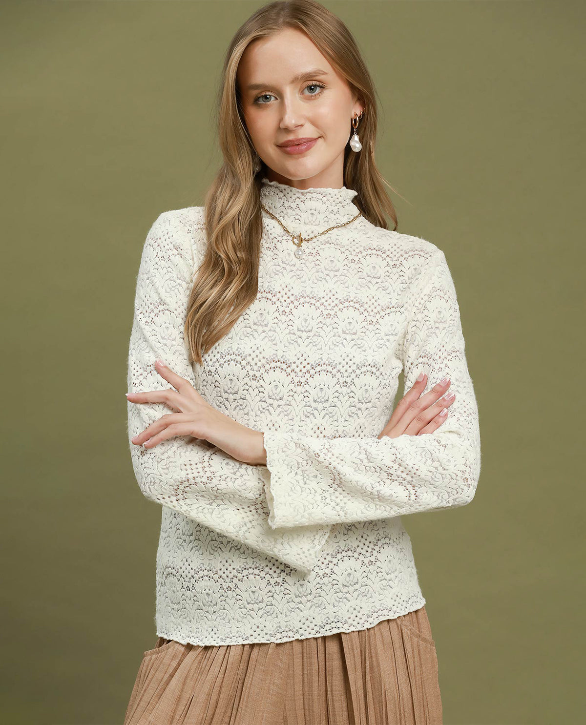 A woman wearing a cream-colored, long-sleeve lace mock neck sweater and a pleated tan skirt, standing against a solid olive green background.