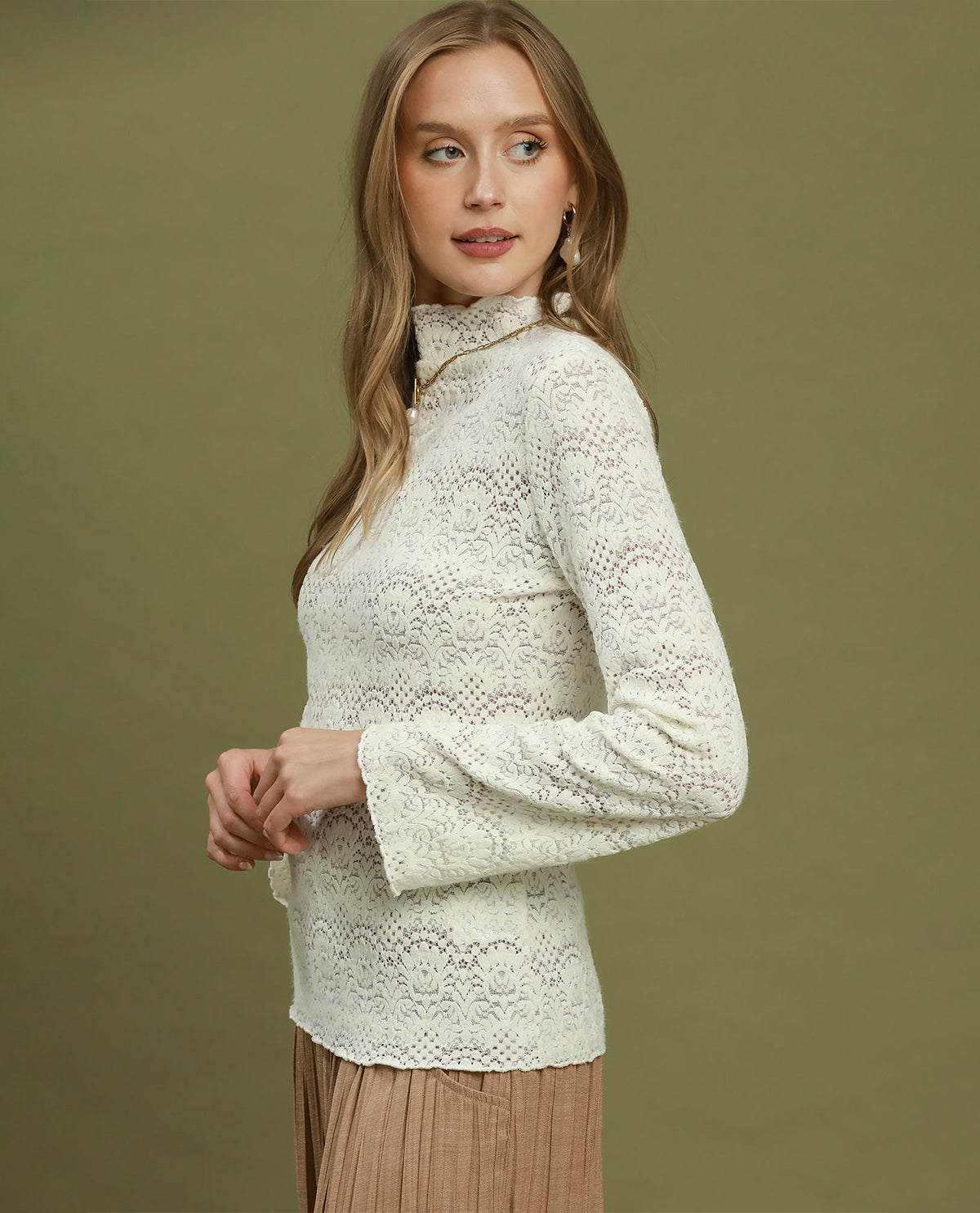 A woman modeling a cream-colored, long-sleeve lace mock neck top against a solid olive green background.