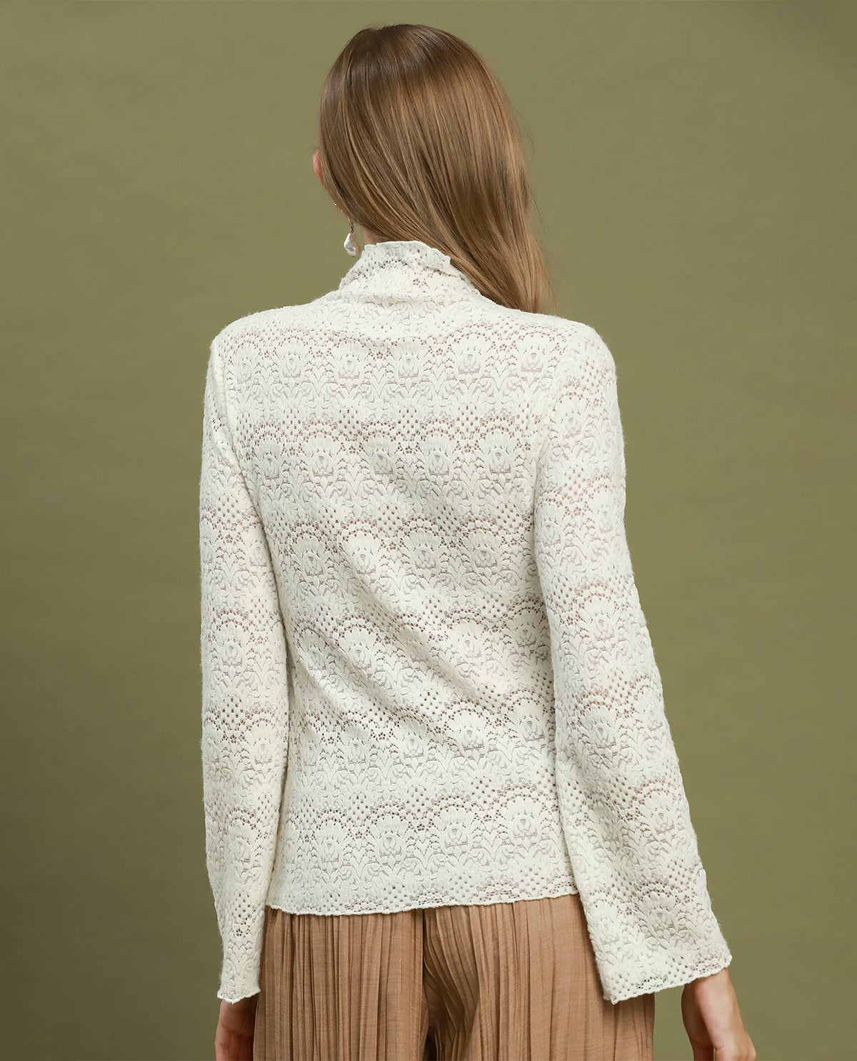 A woman shown from the back wearing a cream-colored, long-sleeve lace knit mock neck top and pleated tan trousers, set against a solid olive green background.