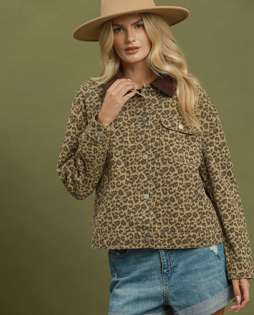 A woman wearing a cropped leopard-print jacket with a dark brown collar, styled with a tan wide-brim hat and blue denim shorts against an olive green background.