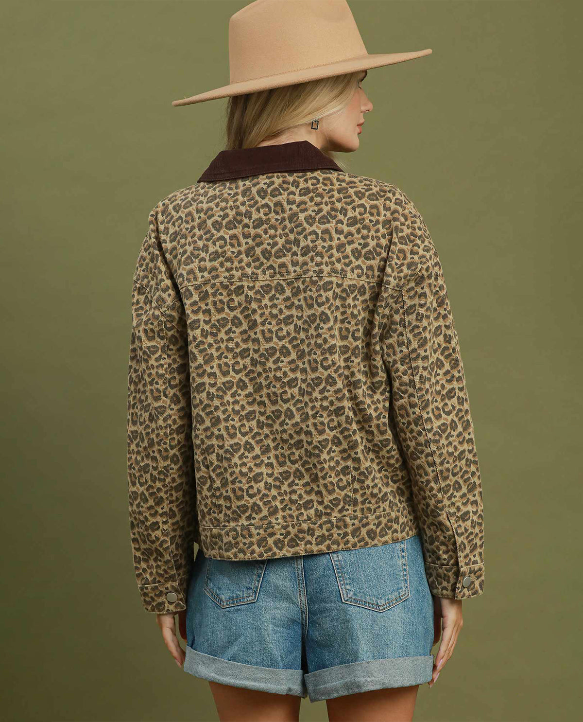 A woman shown from the back wearing a leopard-print long-sleeve shirt jacket with a dark brown corduroy collar, paired with denim shorts and a beige wide-brim hat, against a solid olive green background.