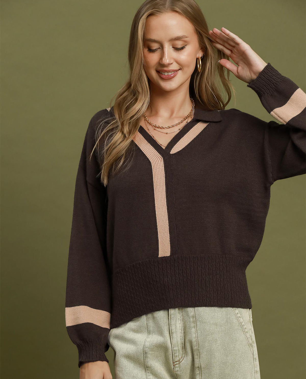 A woman wearing a dark brown knit sweater with beige contrast panels on the collar, front placket, and cuffs, paired with light green jeans, set against a solid olive green background.