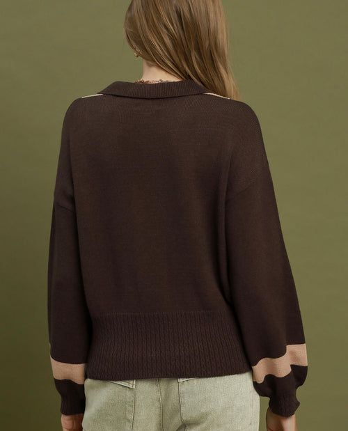 A woman viewed from the back wearing a dark brown knit sweater with beige contrast panels at the collar, shoulders, and cuffs, paired with light olive pants against a solid olive green background.