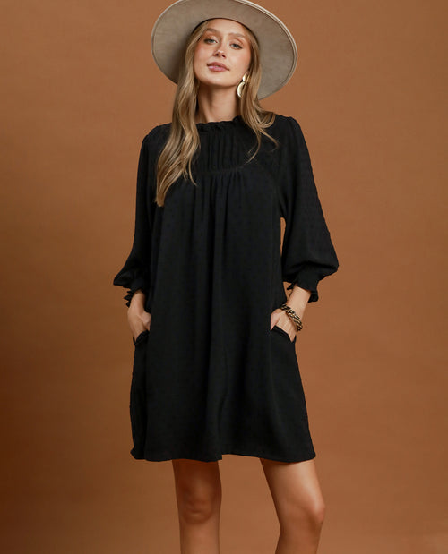 A woman modeling a loose black mini dress with three-quarter sleeves and side pockets, paired with a wide-brimmed beige hat, against a solid warm brown studio background.