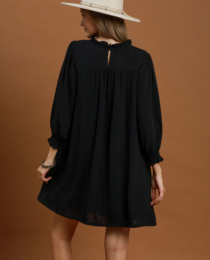A woman wearing a loose black long-sleeve dress with a gathered neckline and puff sleeves, shown from the back against a solid warm brown studio background. She is also wearing a light beige wide-brim hat.