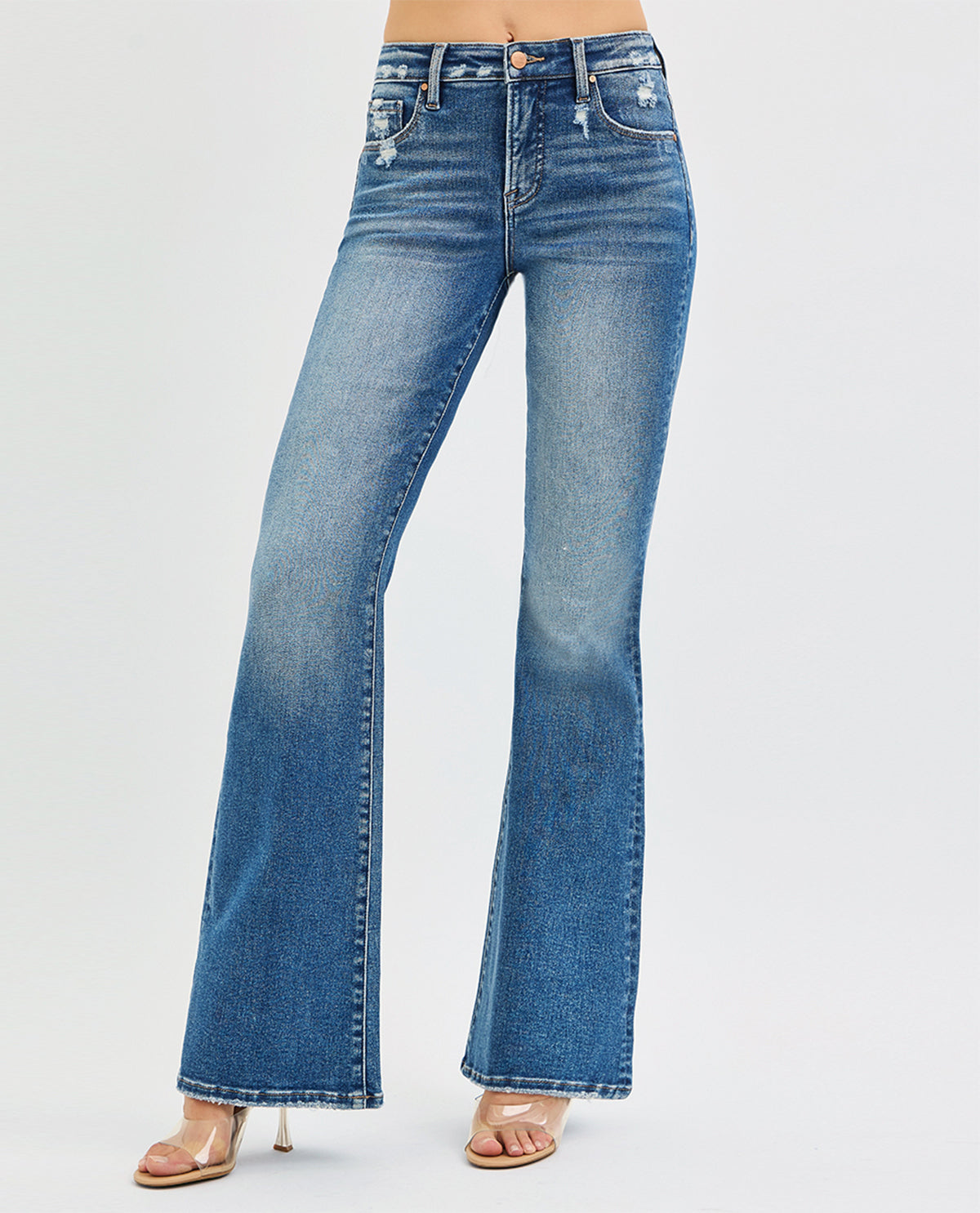 A model from the waist down wearing medium-wash blue denim flare jeans with distressed details and clear high-heeled sandals, set against a plain light gray studio background.