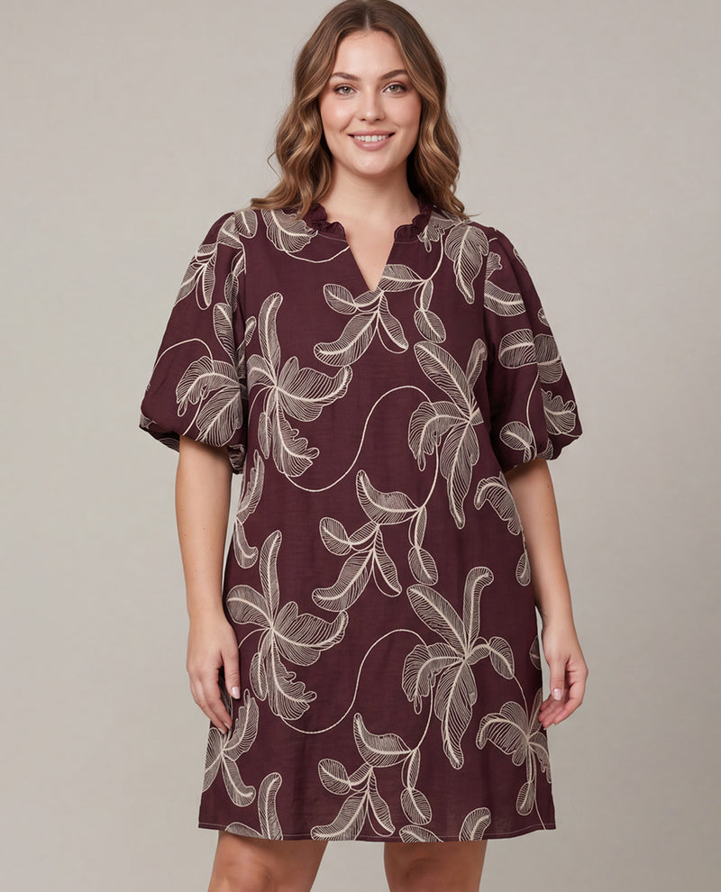 A woman modeling a short-sleeve V-neck dress with a dark burgundy base and a light beige floral vine pattern, set against a solid light gray studio background.