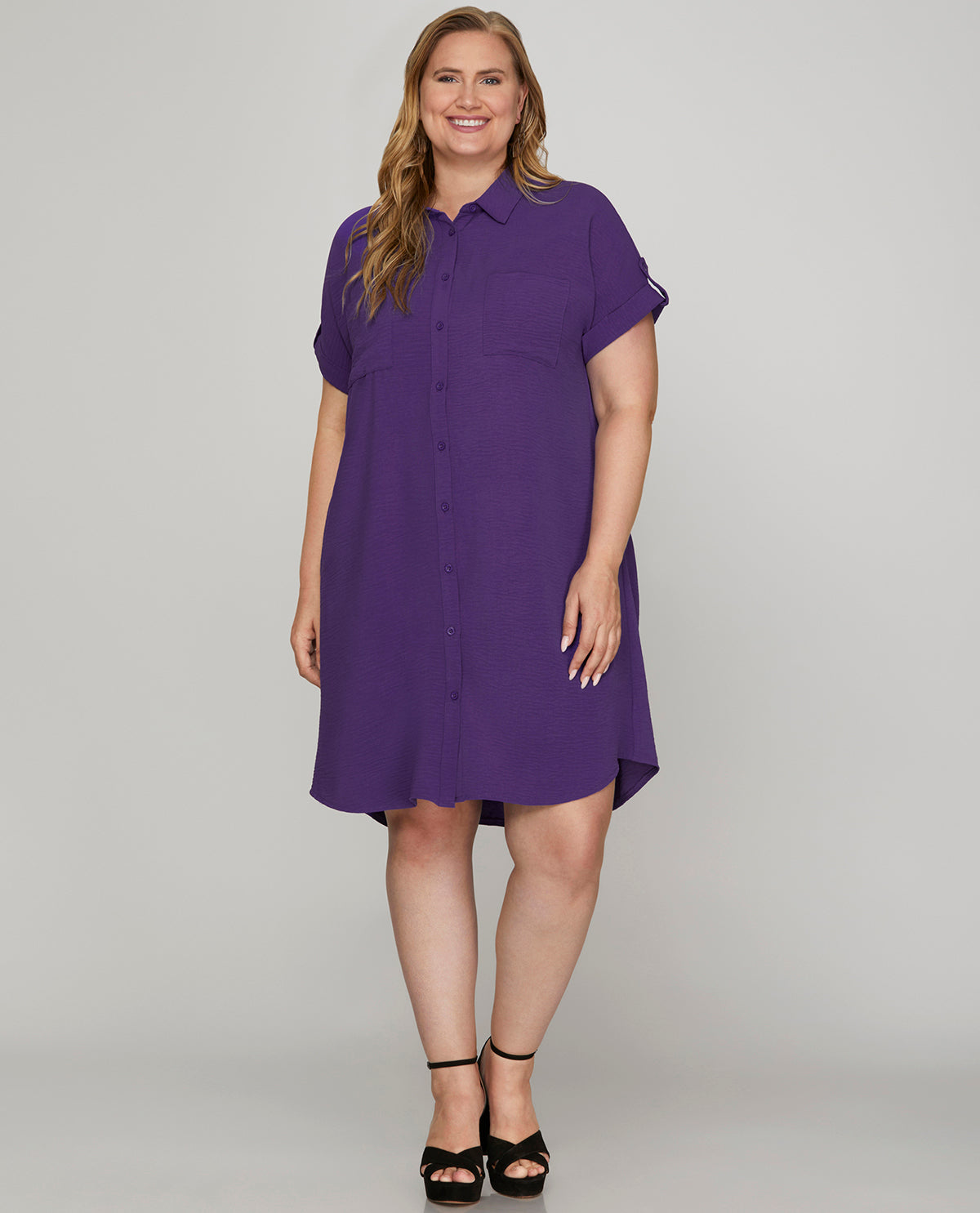 A woman modeling a purple short-sleeve button-up shirt dress with a relaxed, oversized fit and a slight A-line silhouette, paired with black strappy high-heeled sandals against a light gray studio background.