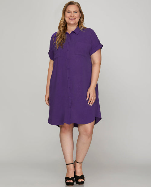 A woman modeling a purple short-sleeve button-up shirt dress with a relaxed, oversized fit and a slight A-line silhouette, paired with black strappy high-heeled sandals against a light gray studio background.