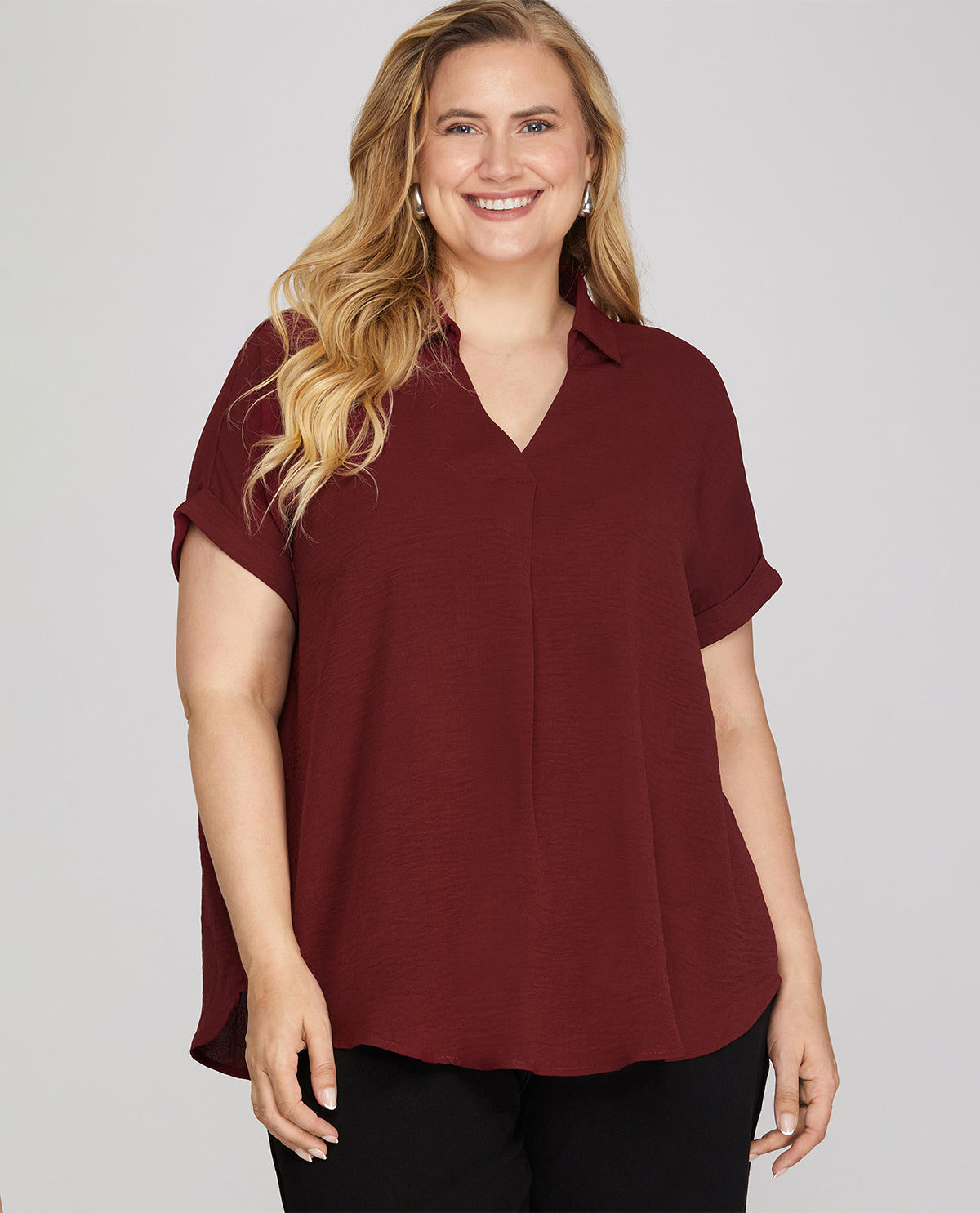 A woman modeling a short-sleeve collared blouse in deep burgundy, paired with black pants, against a solid light gray studio background.