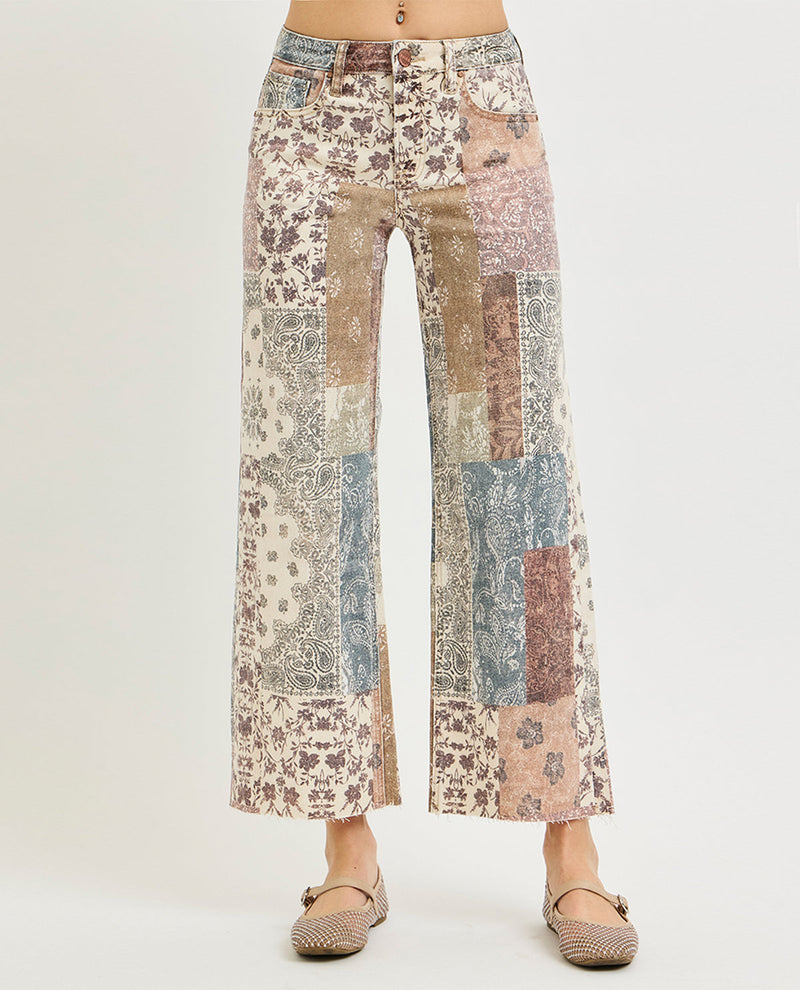 A pair of wide-leg cropped pants with a multicolor patchwork print of vintage floral and paisley patterns in beige, blue, pink, and brown, shown on a model from the waist down wearing flat shoes against a light gray background.