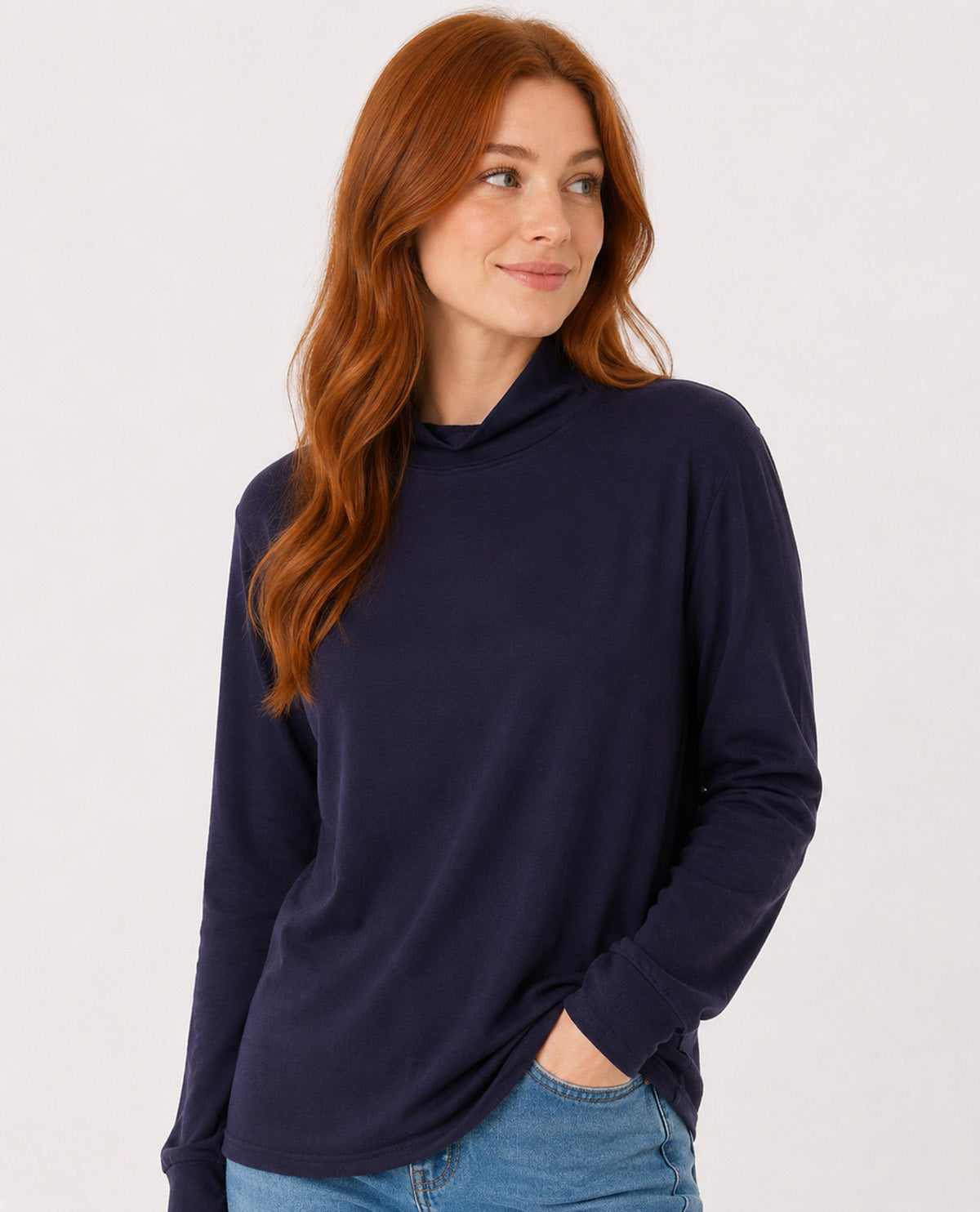 A woman with long red hair wearing a dark navy blue long-sleeve mock neck top and blue jeans, standing against a plain light gray background.