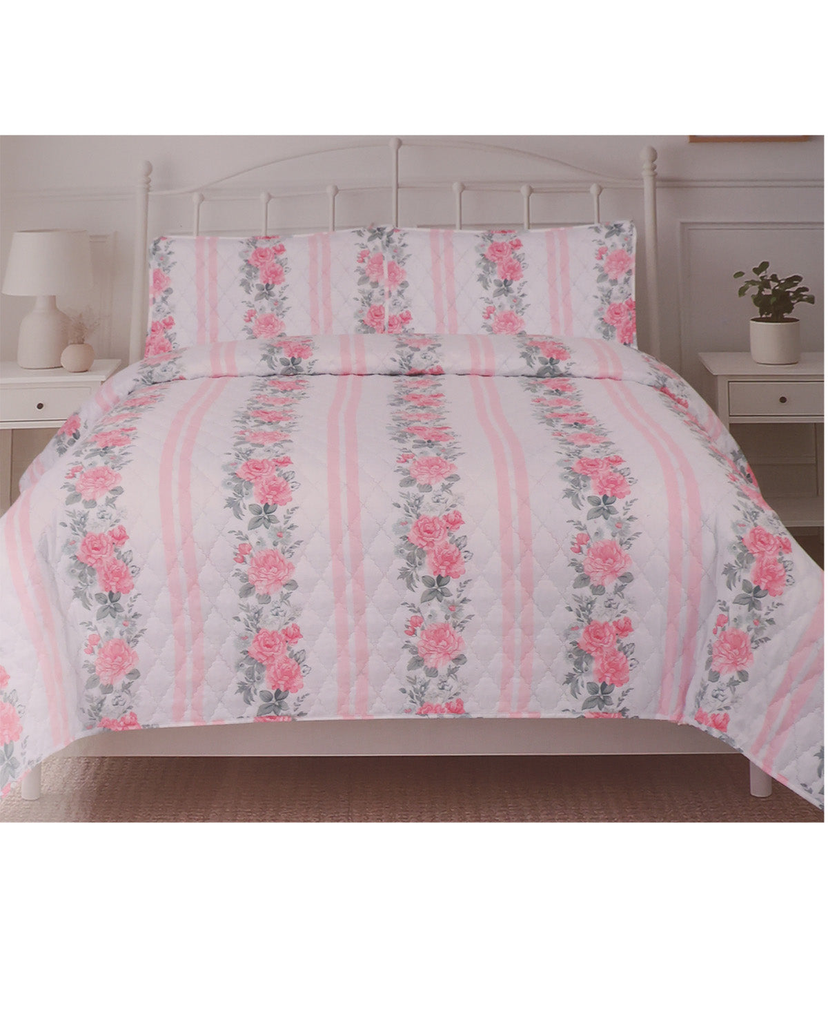 A pink and white floral striped quilted comforter set with matching pillowcases on a white metal bed frame, flanked by white bedside tables with lamps and a small plant.