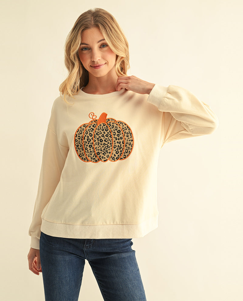 A woman modeling a cream-colored crewneck sweatshirt with a leopard-print pumpkin graphic on the front, paired with blue jeans, against a solid light beige background.