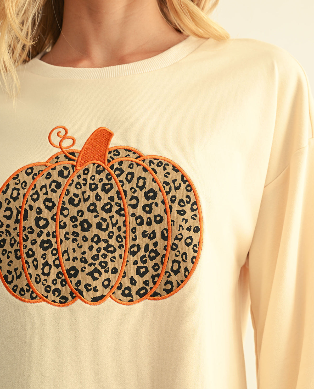 A close-up of a person wearing a cream-colored crewneck sweatshirt with a large leopard-print pumpkin applique on the front.