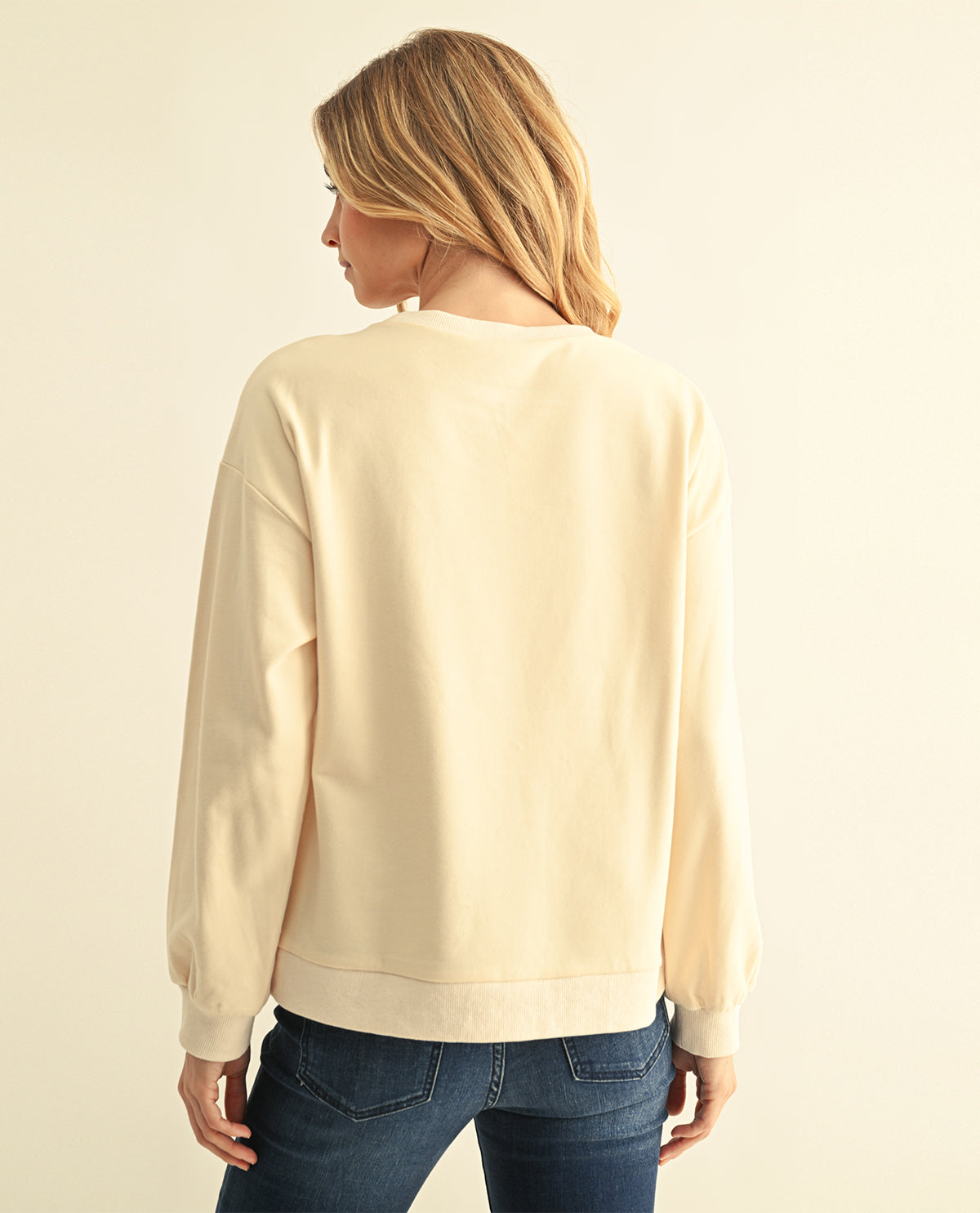 A woman viewed from the back wearing a cream-colored crewneck sweatshirt and dark blue jeans, set against a plain light beige background.
