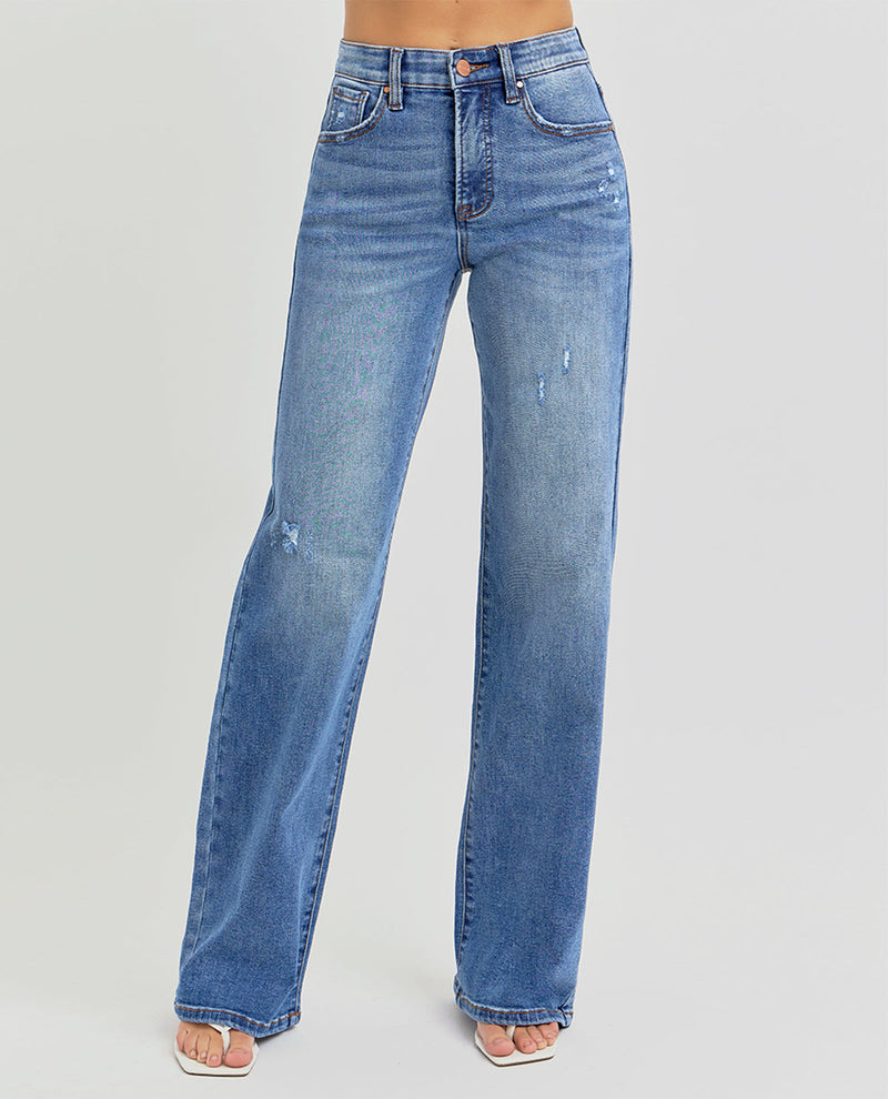 A person wearing medium-wash blue denim jeans with a straight-to-wide leg fit, visible distressing, and a high waist, paired with white slide sandals against a light gray studio background.