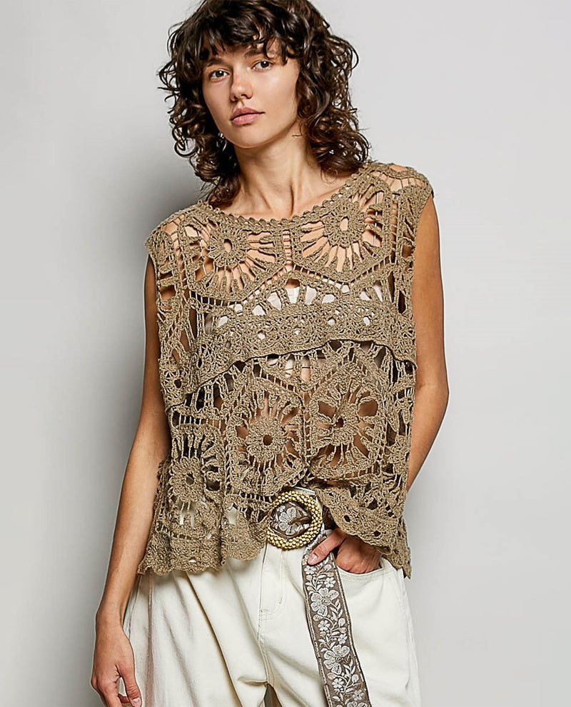 A woman wearing a sleeveless, open-knit crochet top in a taupe-brown color, paired with cream pants and a patterned belt, against a plain light gray studio background.