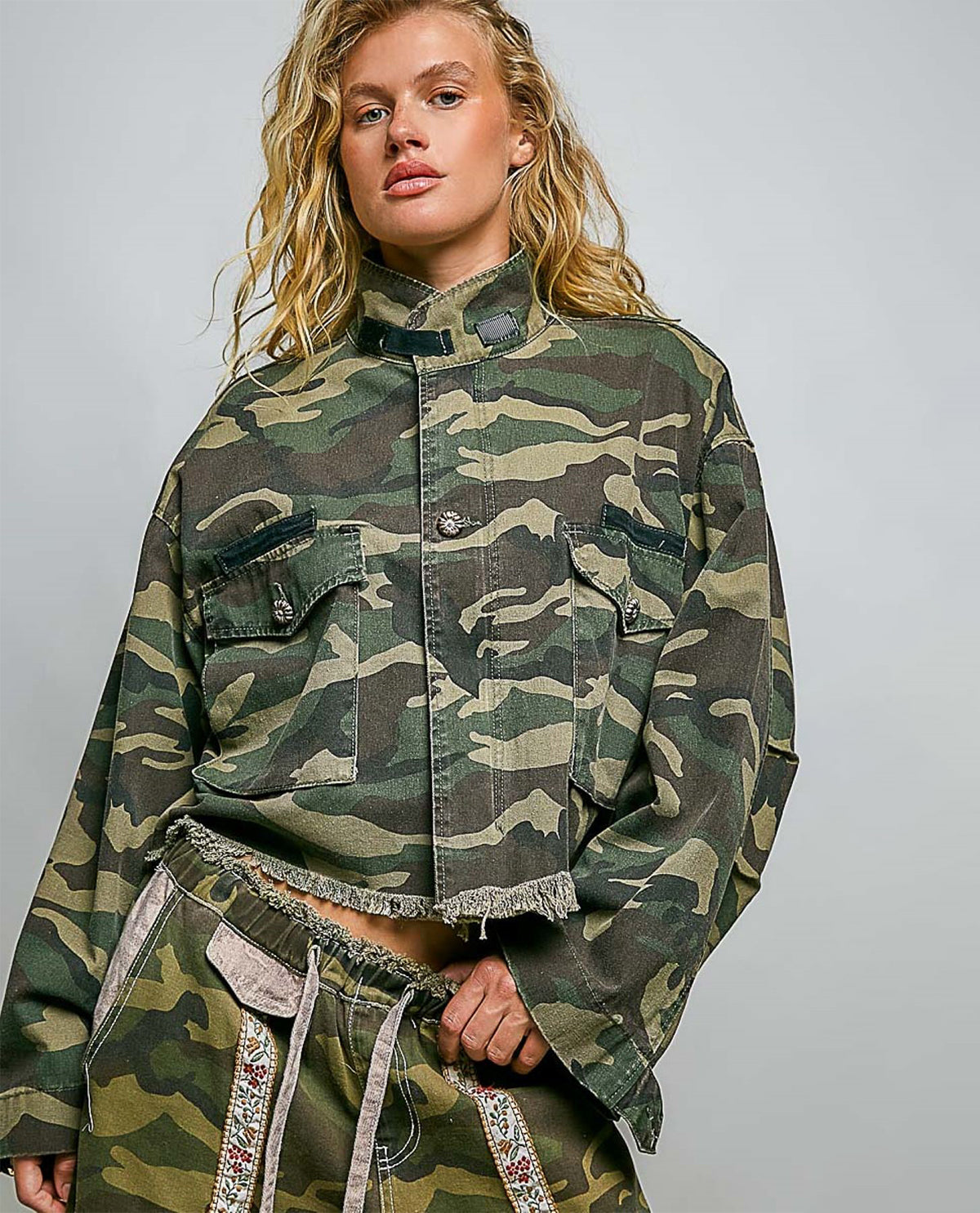 A model wearing a cropped, long-sleeve camouflage jacket with a high collar and frayed hem, paired with matching camouflage pants, against a plain light gray background.