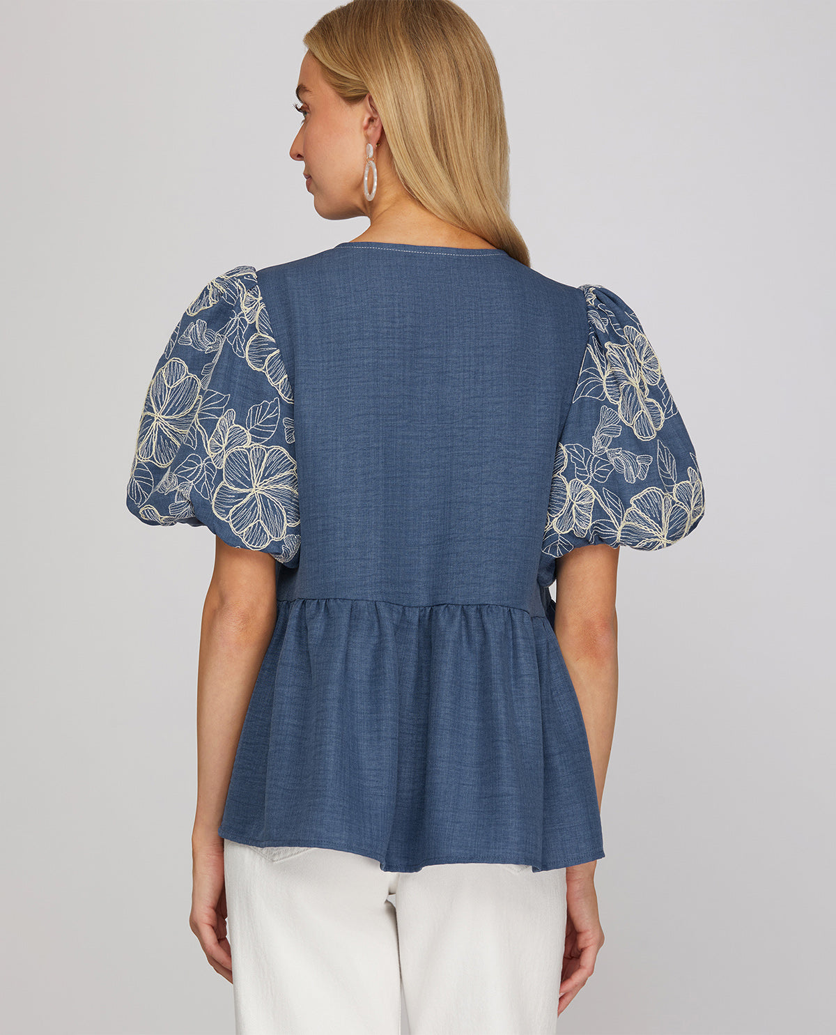 A woman shown from the back wearing a blue peplum top with white floral lace puff sleeves, paired with white pants, against a light gray studio background.
