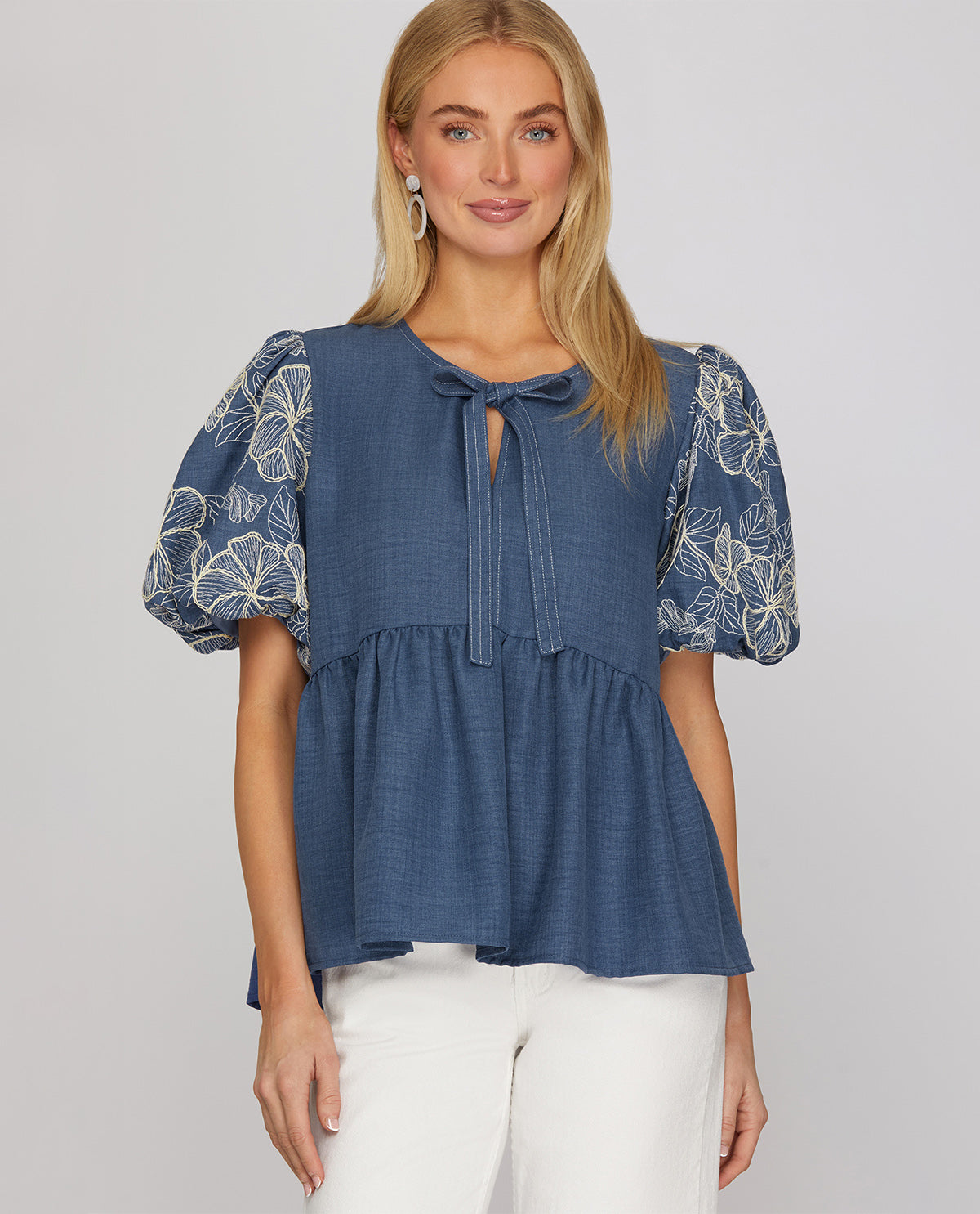 A woman modeling a blue tiered blouse with short puff sleeves featuring white floral embroidery, a tie-neck detail, and a keyhole cutout, paired with white pants against a light gray studio background.