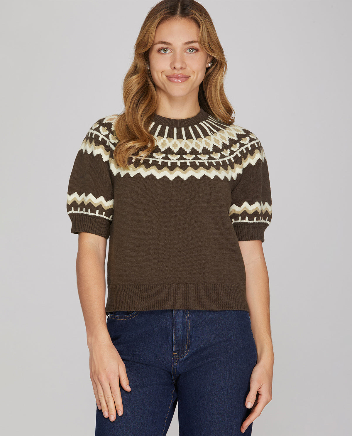 A woman wearing a brown short-sleeve knit sweater with a cream and brown geometric Fair Isle pattern across the yoke and sleeves, paired with dark blue jeans, against a solid gray studio background.