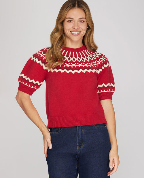 A woman wearing a red short-sleeve knit sweater with a cream Nordic Fair Isle pattern across the chest and sleeves, paired with dark blue jeans, against a solid light gray background.