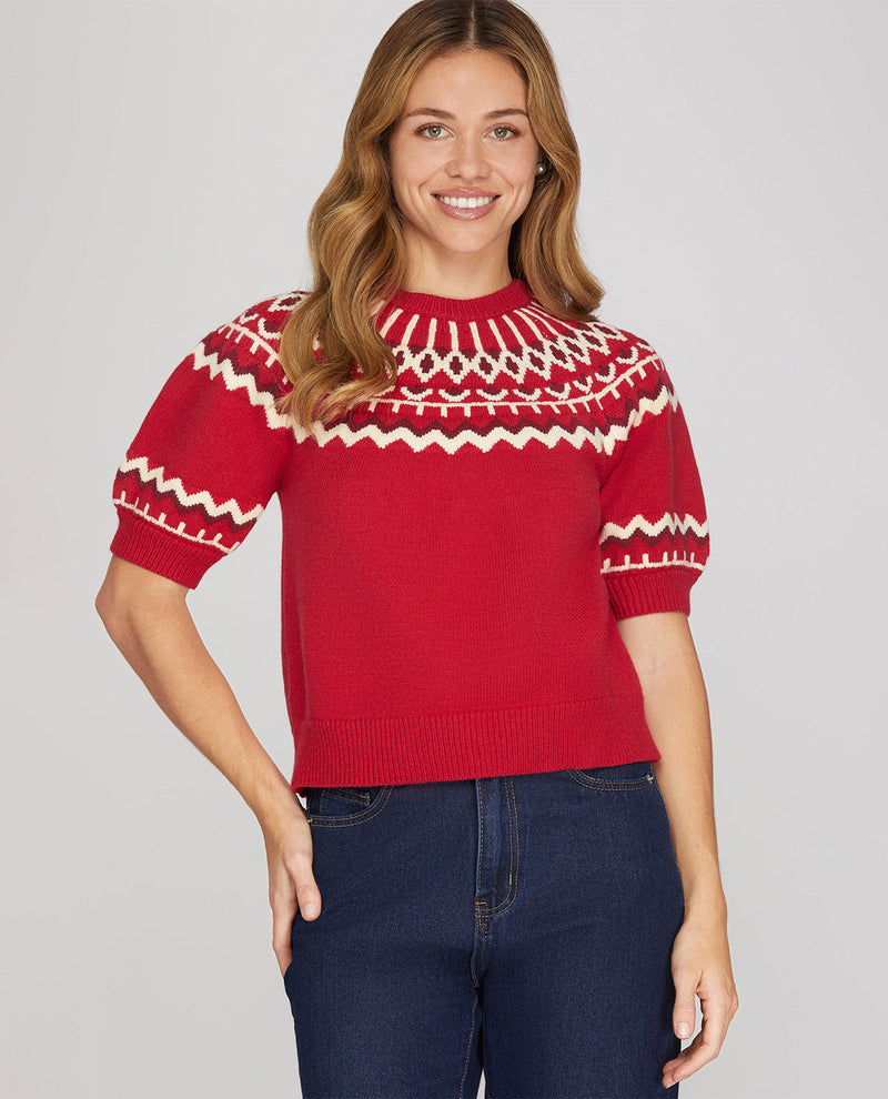 A woman wearing a red short-sleeve knit sweater with a cream Nordic Fair Isle pattern across the chest and sleeves, paired with dark blue jeans, against a solid light gray background.