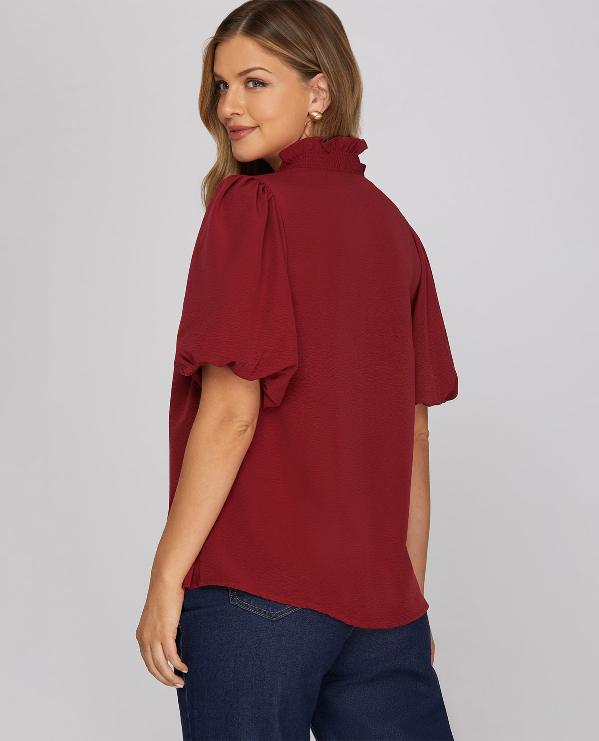 A woman wearing a dark red short-sleeve blouse with ruffled trim and dark blue jeans, photographed from the back against a light gray studio background.