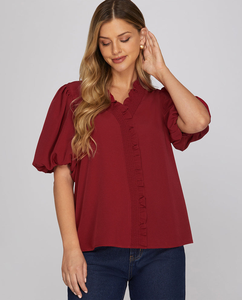 A woman wearing a burgundy short-sleeve blouse with puff sleeves and a ruffled placket, paired with dark blue jeans, against a solid gray studio background.