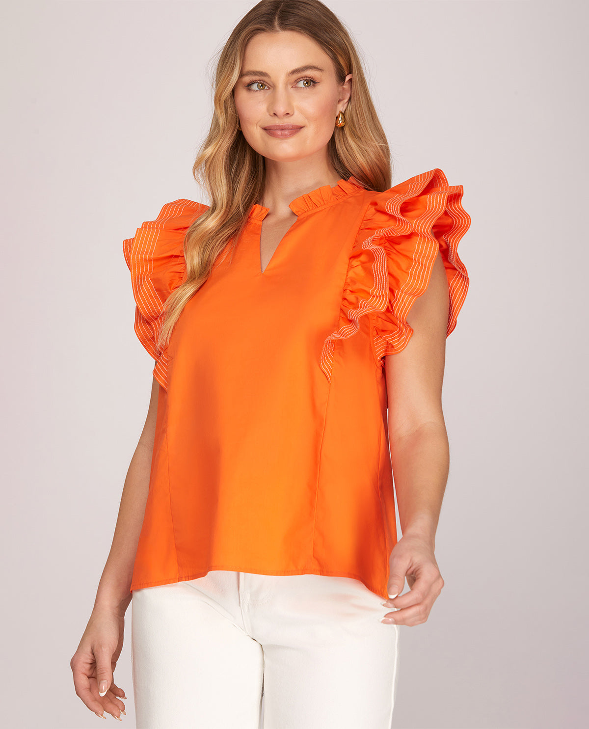 A woman modeling a bright orange sleeveless blouse with ruffled cap sleeves and a split neckline, paired with white pants, against a light gray studio background.