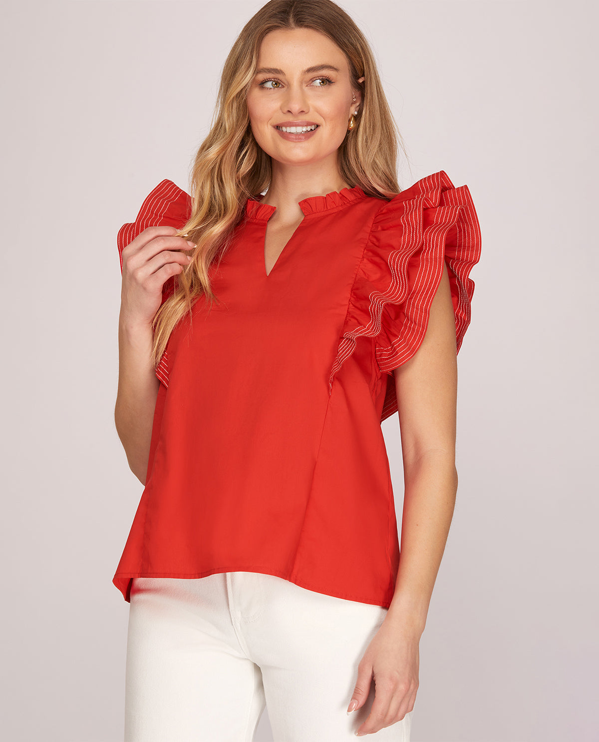 A woman wearing a bright red short-sleeve blouse with ruffled details on the shoulders and a split neckline, paired with white pants, against a light gray studio background.