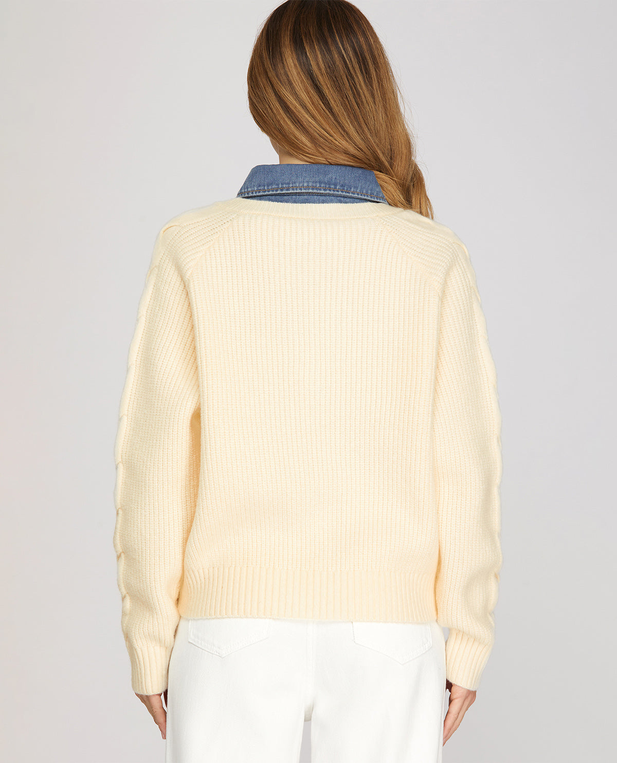 A woman viewed from the back wearing a cream ribbed knit sweater with a blue denim collar, paired with white pants, against a light gray studio background.
