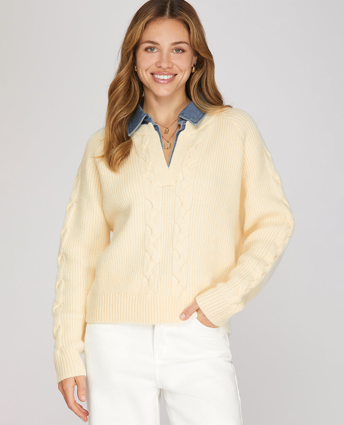A woman wearing a cream-colored cable-knit sweater over a blue denim shirt collar, paired with white pants, against a solid light gray background.