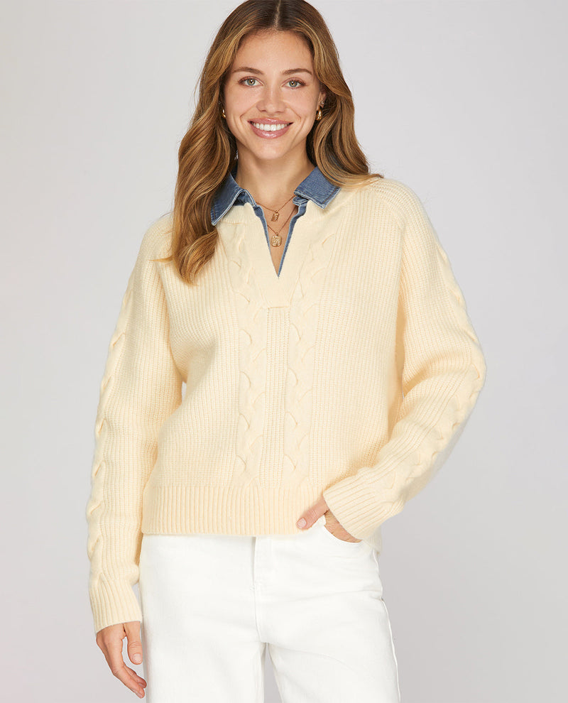 A woman wearing a cream-colored cable-knit sweater over a blue denim shirt collar, paired with white pants, against a solid light gray background.