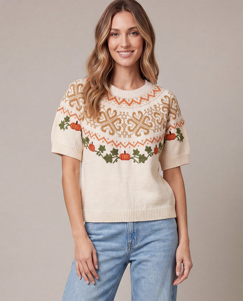A woman wearing a cream-colored short-sleeve knit sweater with a festive autumn pattern of orange pumpkins, green holly leaves, and brown snowflake motifs, paired with light blue jeans, set against a solid beige studio background.