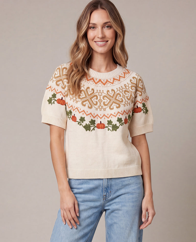 A woman wearing a cream-colored short-sleeve knit sweater with a festive autumn pattern of orange pumpkins, green holly leaves, and brown snowflake motifs, paired with light blue jeans, set against a solid beige studio background.